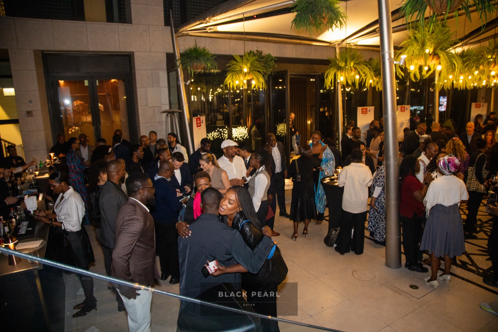 Guests warmly embracing and greeting one another at a Black Pearl evening under string-lit palms